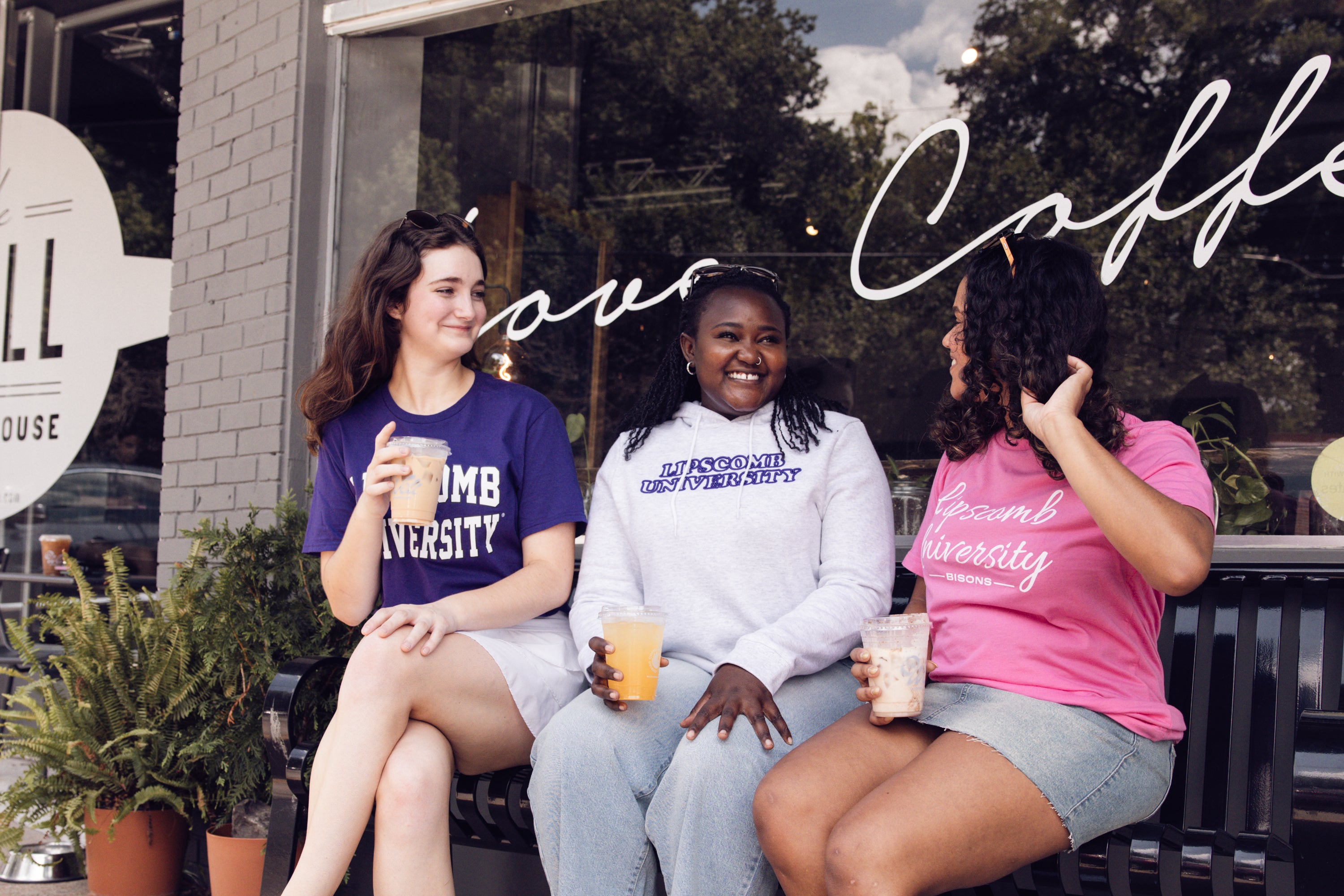 Three women sitting outside a coffee shop, wearing 'Dumb University' shirts.